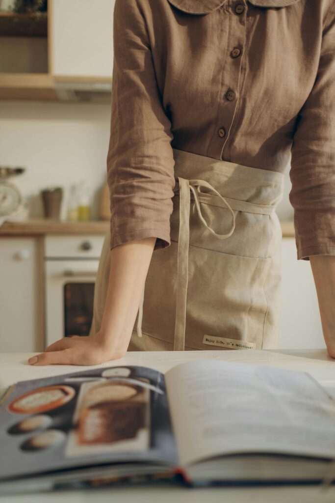 a woman wearing a a brown shirt and a cream apron is leaning on a kitchen counter reading a cook book