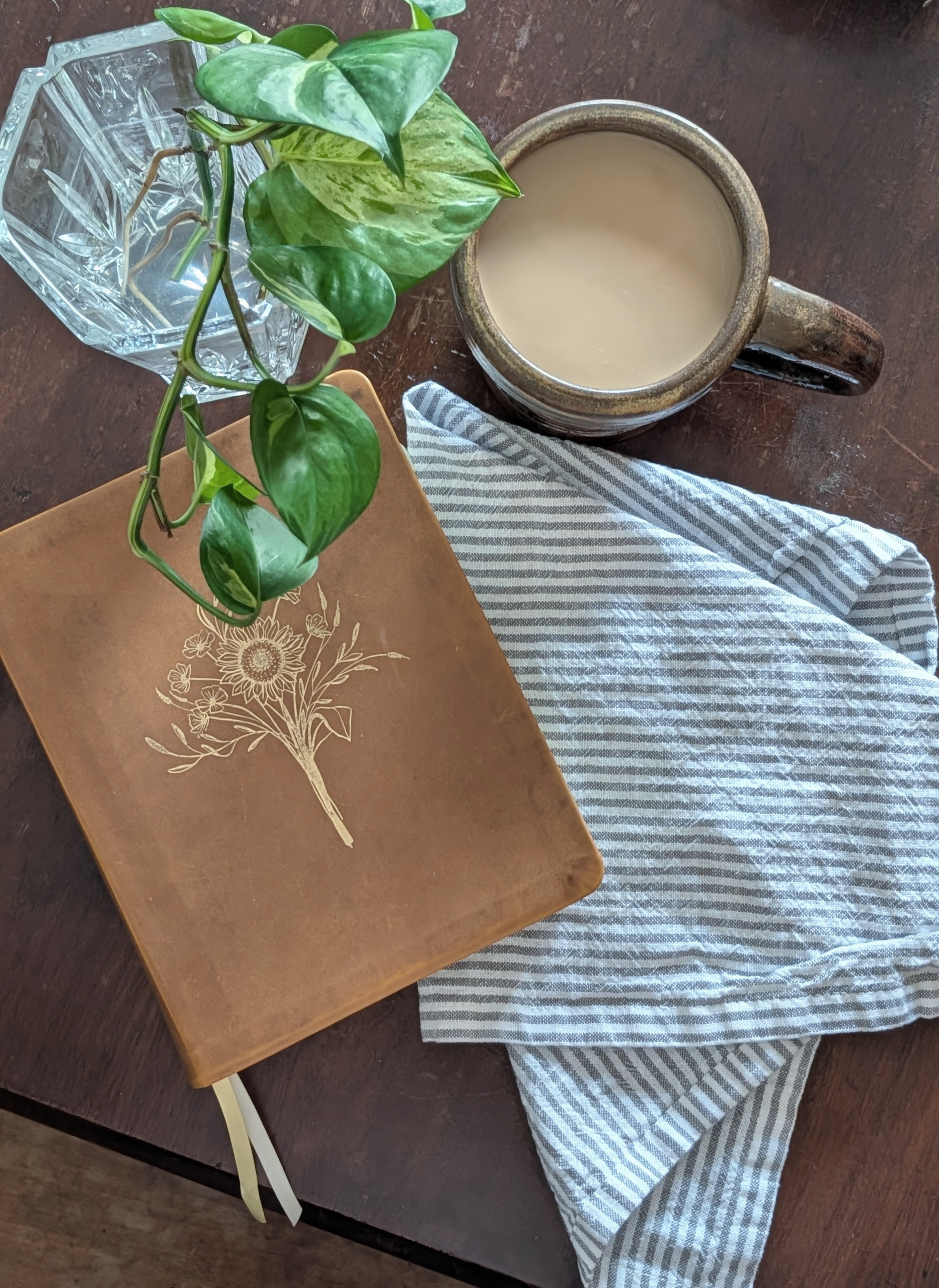 Brown leather Bible, green plant, and a warm coffee mug resting on a striped linen napkin sitting on a dark brown wooden table