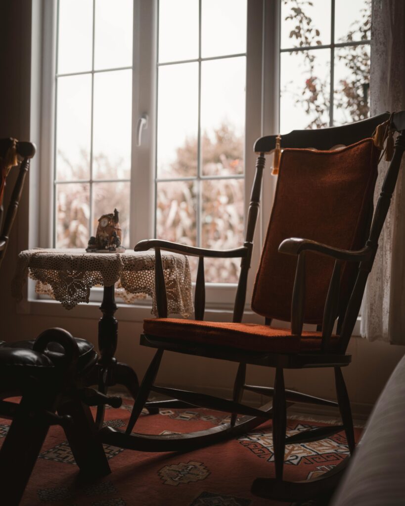 A wooden rocking chair sitting in front of a big window and a small round table nearby