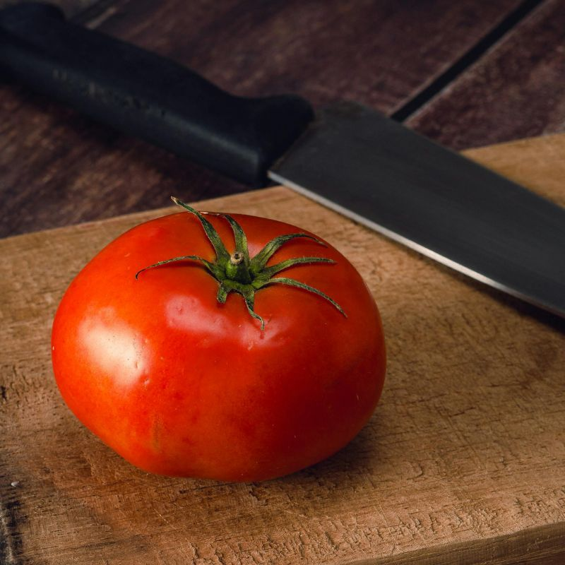 a red tomato sitting beside a black handle chef's knife on a wooden cutting board
